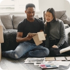 Couple sit on the floor and review paint color samples together while surrounded by moving boxes. 